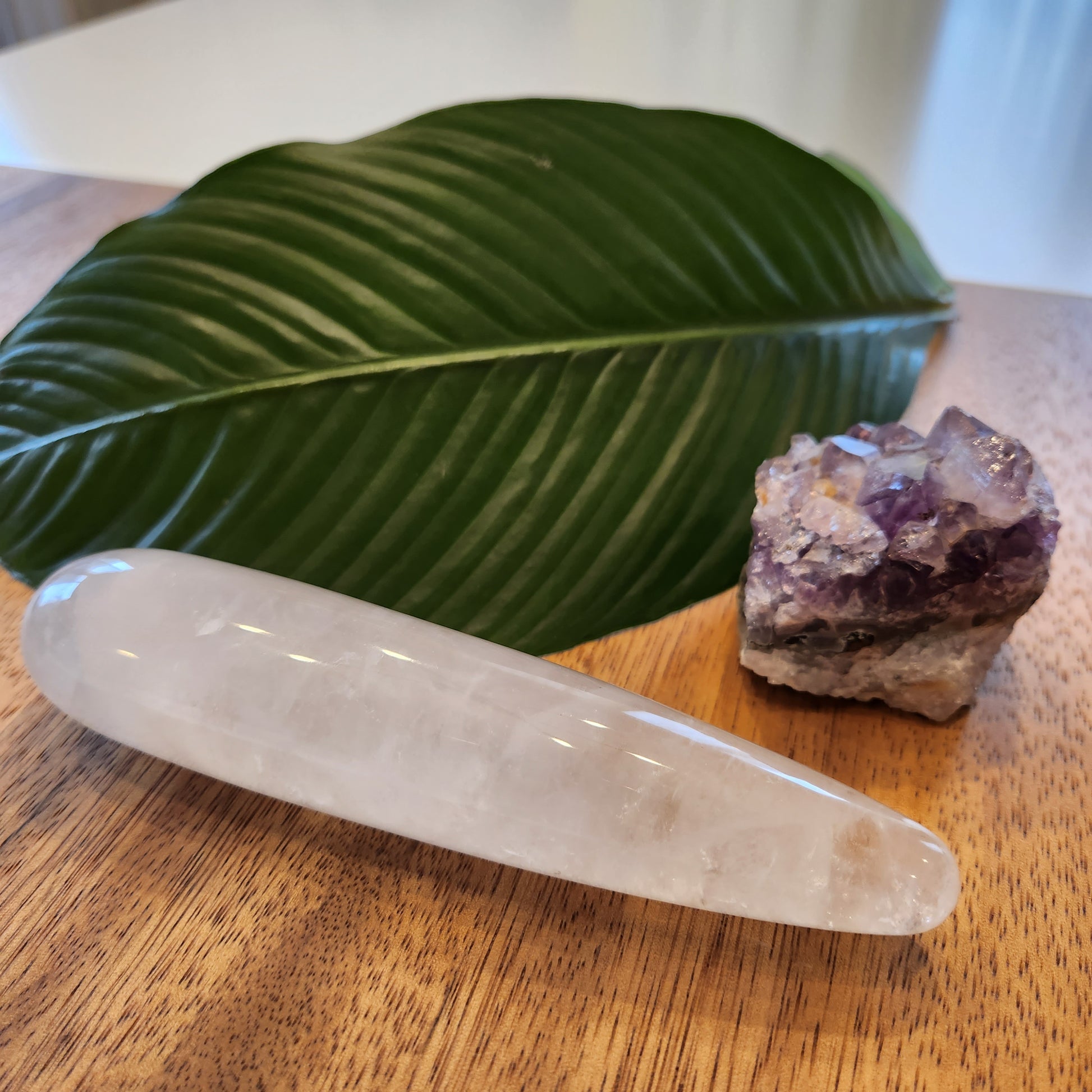 Thick, polished clear quartz crystal wand with a large diameter perfect for sex anal, lying on a wooden surface next to a large green leaf and a rough amethyst geode.