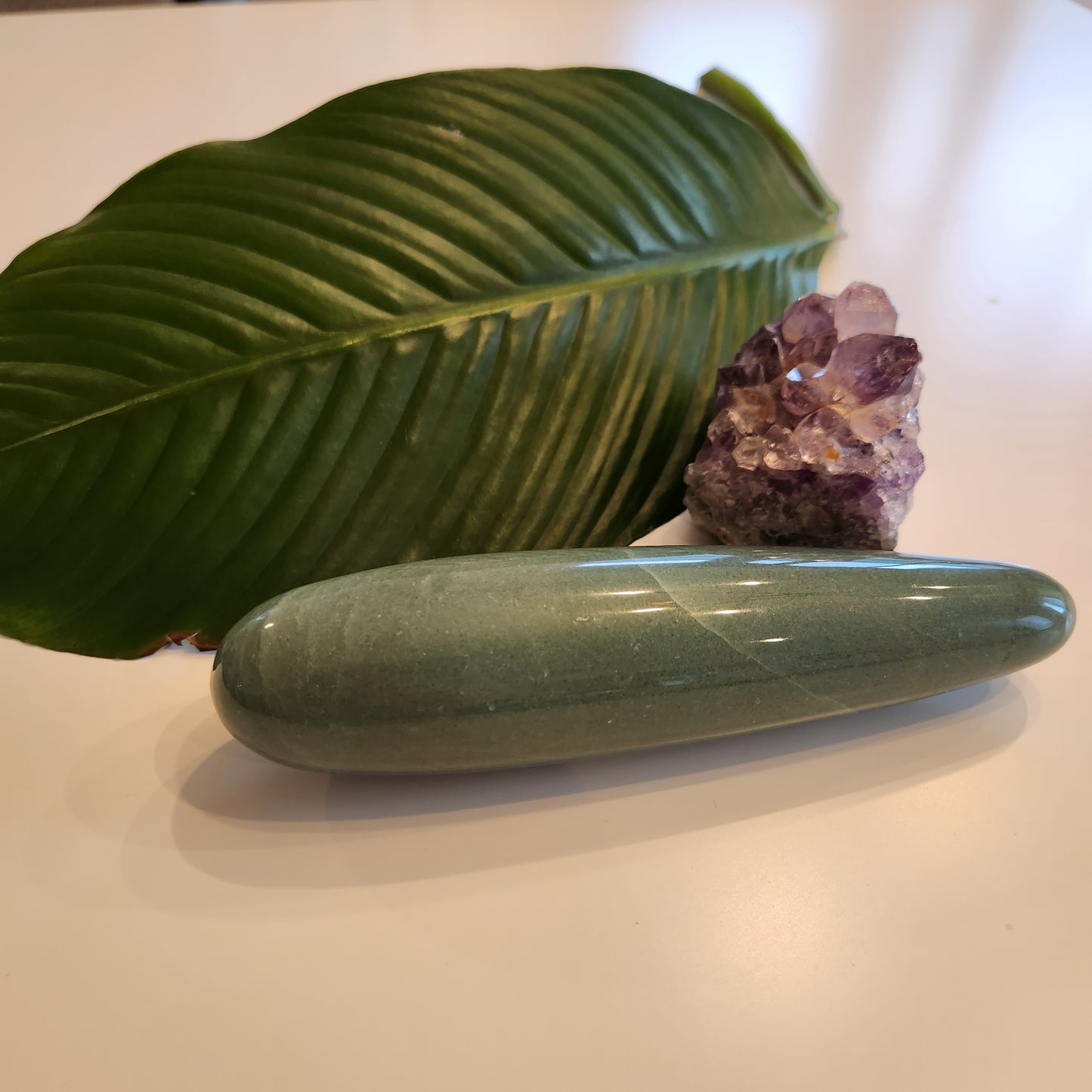 Thick, polished green aventurine crystal wand with a large diameter, lying on a white surface next to a large green leaf and a rough amethyst geode.