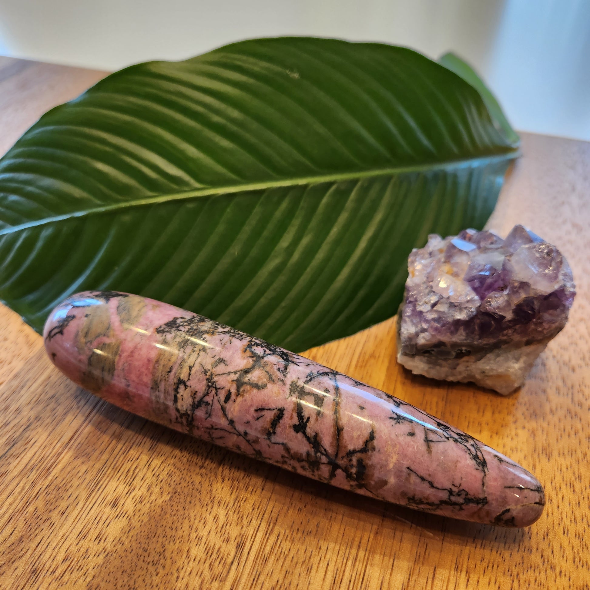 Thick, polished Rhodonite crystal wand with pink and black veins perfect for anal stimulation, lying on a wooden surface next to a large green leaf and a rough amethyst geode.