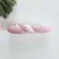 Polished rose quartz yoni wand with a twisted, spiral body and a pointed tip, lying on a white surface with pine needles.