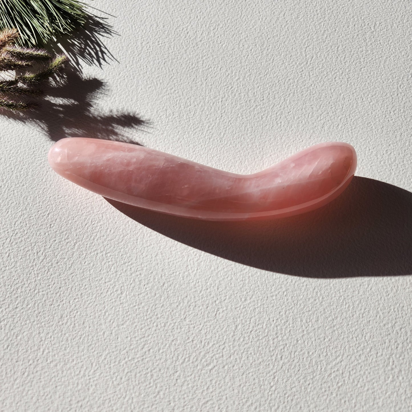 A hand-polished, ergonomically sculpted rose quartz intimate massager lying on a light-colored surface. The curved shape is designed for targeted pleasure