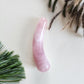 A hand-polished, mini curved rose quartz crystal wand lying on a light-colored surface with a green pine branch and dried grass in the background.