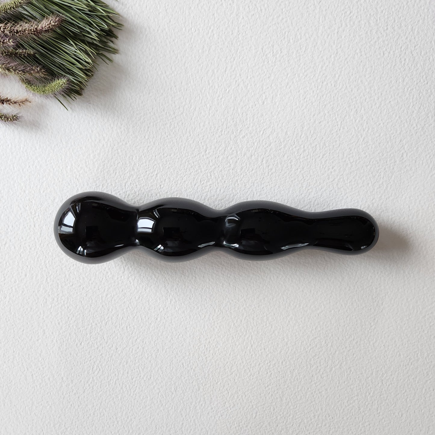 Polished black obsidian yoni wand with a distinct multi-curve or knotted shape, perfect for anal stimulation, lying on a white surface with a sprig of dried foliage.