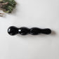 Polished black obsidian yoni wand with a distinct multi-curve or knotted shape, perfect for anal stimulation, lying on a white surface with a sprig of dried foliage.