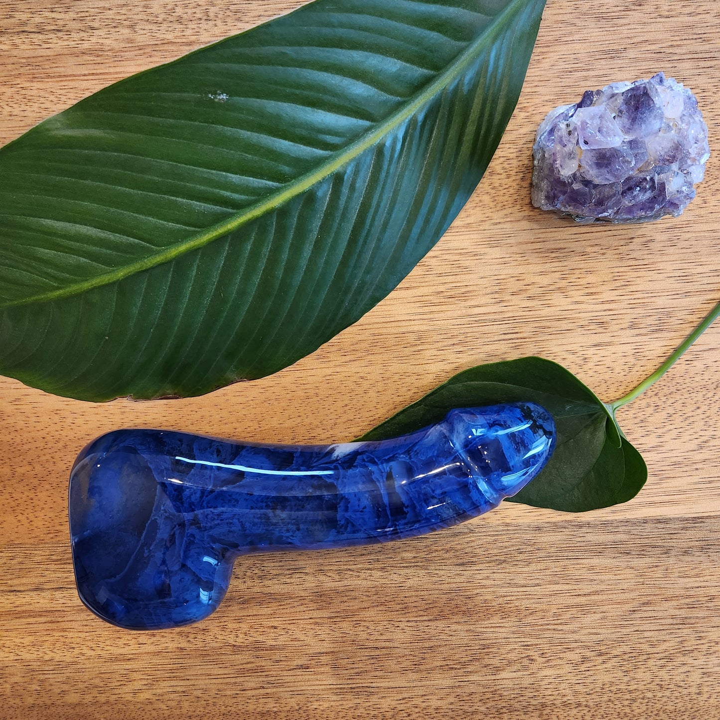 Polished blue crystal wand in a phallic shape with a flowing or "melting" pattern perfect to reach the G-Spot, lying on a wooden surface next to a green leaf and a rough amethyst geode.