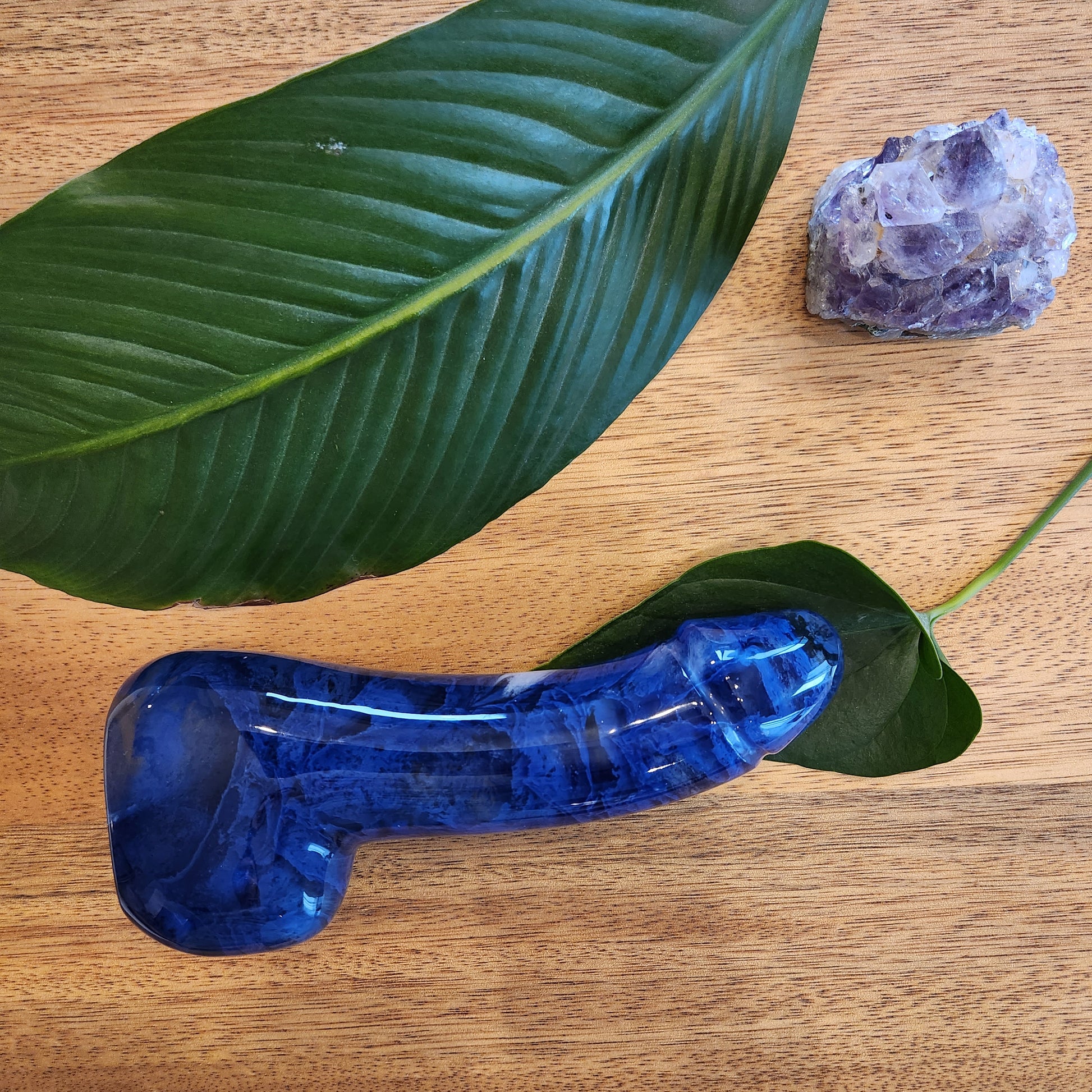 Polished blue crystal wand in a phallic shape with a flowing or "melting" pattern perfect to reach the G-Spot, lying on a wooden surface next to a green leaf and a rough amethyst geode.