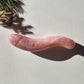 Polished rose quartz wand with a phallic shape and a distinct S-curve, lying on a white surface with pine needles and a strong shadow.